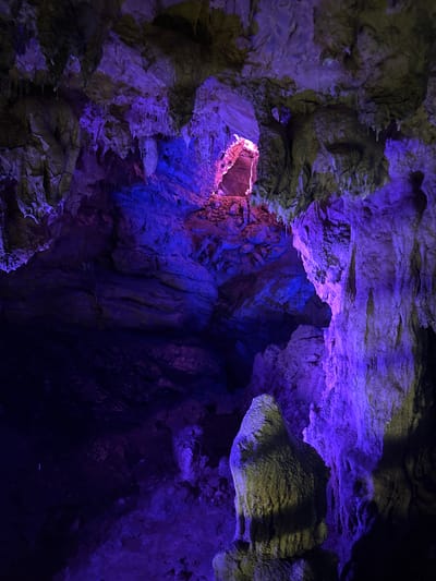 The image captures the interior of a cave, likely a tourist attraction in Sanguansi, China, illuminated with dramatic purple and pink lighting. The cave walls are rough and uneven, featuring various rock formations, stalactites hanging from the ceiling, and stalagmites rising from the floor. The artificial lighting creates a surreal atmosphere, casting deep shadows and highlighting the textures of the rock. There are no people or animals visible in the scene. The environment appears natural, albeit enhanced by artificial illumination, suggesting a managed or developed cave system.
