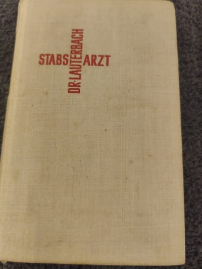 The image shows the front cover of a beige book with red text. The text reads "STABS DR. LAUTERBACH ARZT". The book appears to be aged, with some faint discoloration on the cover. The book is resting on a dark, textured surface that resembles carpet. There are no people or other objects visible in the image. The location in Schwäbisch Gmünd, Germany is not discernible from the image.