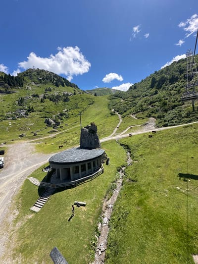 The image depicts a summer alpine landscape in the Plaine Dranse area of Châtel, France, observed from an elevated perspective, likely from a ski lift. The scene is dominated by a vibrant green, grassy valley under a clear blue sky with scattered white cumulus clouds.

In the mid-ground, a distinctive, modern-style chapel with a curved, dark slate roof and multiple windows is centrally located on a grassy slope. A large, natural rock formation, topped with a slender cross, rises vertically immediately behind the chapel. A graveled dirt road runs horizontally across the frame, passing by the chapel; a white recreational vehicle is parked on this road to the left of the chapel. A single individual, dressed in dark shorts and a light top, stands on the green grass directly in front of the chapel, positioned to the left of a dry, rocky stream bed that traverses the slope.

Scattered across the grassy hillsides are several domestic animals: multiple dark-colored bovines (cows) are visible grazing, primarily to the right of the stream, and at least two light-colored equines are present further up the dirt road near the chapel. The surrounding steep mountain slopes are covered in lush green grass, with coniferous trees on the higher elevations, particularly on the peak to the left. Multiple ski lift towers and lines ascend the mountains, with a prominent tower visible on the far right edge of the frame. Numerous narrow dirt paths snake across the distant hillsides. The sun casts distinct shadows, indicating daytime conditions.