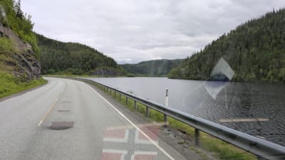 This is a roadside view in Mosjøen, Norway, likely captured from a vehicle. The scene depicts a winding asphalt road curving to the left, bordered by a metal guardrail and a rocky, tree-covered embankment on the left. To the right of the road is a body of dark, rippling water, likely a fjord or lake, with heavily forested hills rising in the distance. The sky is overcast with a muted grey hue. The time of day appears to be daytime, given the ambient light, but the cloudy conditions suggest it could be morning, afternoon, or evening.
There are no visible people or clear objects in the immediate foreground. However, the surface of the water has a faint, superimposed image of a Norwegian flag, indicated by the red and white cross pattern. This reflection or overlay suggests a possible connection to Norway or a deliberate visual element added to the media. Another superimposed graphic appears as a diamond shape with a dark circle in its center, also reflected on the water. The road surface itself shows several patched areas, indicating wear and tear. The overall mood is somewhat subdued due to the overcast weather and the vast, natural landscape. There is no visible text in the image.
