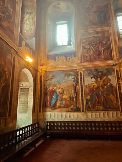 The image captures the interior of a historic building, possibly a church or chapel, characterized by religious paintings and a sense of reverence. The scene is dominated by large murals depicting biblical scenes or religious figures on the walls. A set of wooden benches are present, suggesting a space for prayer or reflection. A decorative wooden rail separates the seating area from an adjacent corridor where a plaque with Cyrillic text can be seen. The architectural details such as arched doorways and window frames, contribute to the building's historical character. The overall atmosphere is dim, with lighting provided by wall-mounted fixtures and natural light entering through a high window. While the location is noted as Fedotovo, Russia, the image itself is the main subject.
