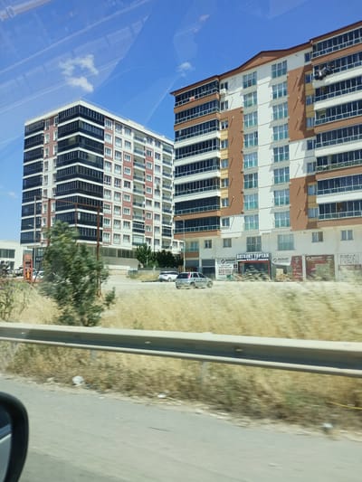 In the city of Yahşihan, Turkey, two modern apartment buildings stand tall under a clear blue sky with scattered clouds. The building on the left is characterized by its white and dark-paneled facade, with numerous windows lining each of its floors. The building on the right features a mix of white and brown panels, with balconies and windows on every level. In the foreground, dry, golden grass and a metal guardrail line a road. Several cars are visible on the road and in front of the buildings. The lower level of the right building has storefronts with signage. The scene is captured from a moving vehicle, indicated by the reflection on the glass and the motion blur in the image.