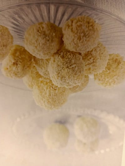A plate of coconut-covered confections sits on a white surface. The confections are round and pale yellow in color, with a rough texture from the shredded coconut coating. They are arranged in a cluster, with some overlapping. The plate itself is clear glass with a fluted edge. The background is blurred and appears to be a light-colored surface, possibly a tablecloth or countertop. There are no people or discernible environmental features to suggest a specific location.