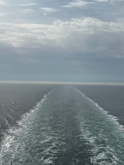 The image depicts a view of the sea, with a ship's wake prominently visible, extending from the bottom center of the frame to the horizon. The sea is moderately choppy, with sunlight reflecting off the wave crests. In the distance, near the horizon, a faint outline of land or a distant ship is discernible. The sky is overcast with clouds, transitioning from brighter, lighter tones towards the left side of the image. The location is at sea, near an unknown coastline in France.