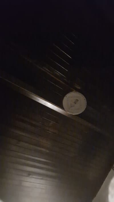 The image captures a low-angle view of a dark, wooden-paneled ceiling with distinct horizontal planking. A circular, light-colored object, possibly a light fixture or a cover, is mounted centrally on the ceiling. The object appears to have some markings or text inscribed on its surface, but these are not clearly legible. The overall lighting is dim, suggesting the photograph was taken in low-light conditions. The environment appears to be indoors. The location of Kosharitsa, Bulgaria is not visually represented in the image itself.