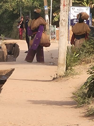 The image displays an outdoor scene on a dirt pathway in San Martín, Colombia, illuminated by bright daylight. In the mid-ground, a central figure walks to the right, wearing an elaborate costume consisting of a dark fabric covering the head and upper body, layered with shaggy light brown material forming a cape around the shoulders and a skirt-like garment at the waist. The arms are covered with patterned purple sleeves, and the legs with matching purple trousers. To the right, partially obscured by a light-colored utility pole, a second costumed figure stands with its back to the viewer, exhibiting similar dark head covering, shaggy light brown shoulder and waist coverings, and a light-colored skirt.
In the left background, an uncostumed woman with dark hair wears a black top and red trousers, walking towards the left. Behind her, a man in dark clothing walks towards the right. The pathway surface is predominantly light brown packed earth, with a concrete slab section over an open drainage ditch in the foreground-left. Various green foliage and shrubs line the path, particularly on the right. A second, thinner utility pole is visible further back, and behind it, a white sign with blue lettering reads "The Centre for Counselling" with a logo, indicating an institutional building nearby. Shadows are visible on the ground, cast by the figures and the utility pole.
