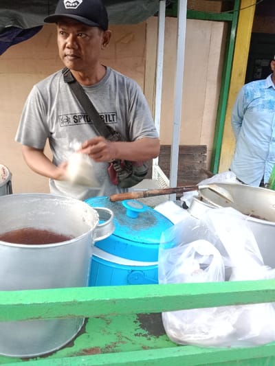 A vendor in a gray t-shirt and black cap stands behind a food stall. He holds a plastic bag. The stall has a large aluminum pot filled with a dark liquid, a blue insulated container, and several clear plastic bags filled with food. A large metal spoon rests on the container. Another man in a light blue shirt is partially visible to the right. The stall is painted green.