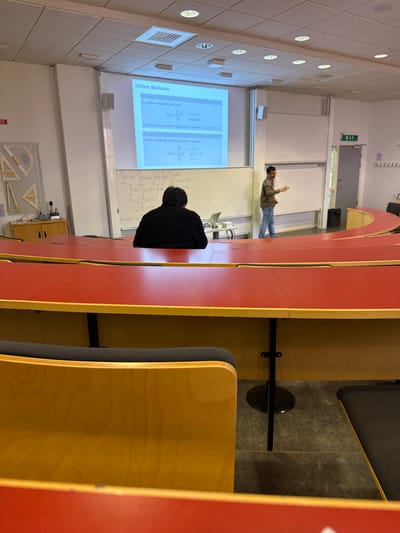 Here's a concise description of the image:
**Setting:** Interior of a lecture hall, likely at Jönköping University in Sweden.
**People:** One man is standing near the whiteboard, lecturing. Another person is seated, presumably a student.
**Objects:** Rows of desks and chairs, a whiteboard displaying information about uniform distribution, a projector screen showing the same information, geometrical tools mounted on the wall.
**Actions:** Lecturer is explaining the uniform distribution concept to the class.
**Interactions:** Lecturer interacting with class to explain uniform distributions.
**Environment:** The lecture hall seems well-lit with rows of fixed seating. The room has standard academic facilities.