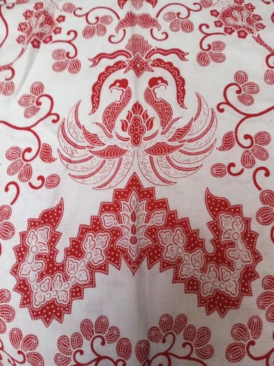 A white fabric is patterned with red traditional Indonesian batik motifs. Prominent designs include two mythical birds facing each other, elaborate floral arrangements, and curling vines with small, round flowers. The pattern is dense and repeated across the fabric, creating a complex and visually engaging textile. The red color pops against the white background.