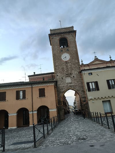 The image depicts a street scene in San Giovanni in Marignano, Italy, under an overcast sky. A central, multi-story brick bell tower with a prominent archway at its base serves as a town gate. The tower features a white clock face with dark hands indicating approximately 1:55, an open belfry with visible bells, and is topped by a weather vane.

To the left of the tower, a two-story building with a light orange-peach facade displays three arched ground-floor entrances and two first-floor windows fitted with closed dark brown shutters. A black scooter is parked on the sidewalk directly in front of this building. To the right of the tower, a two-story building with a light beige facade features two first-floor windows with closed dark brown shutters and a partially visible ground-floor window. Above this building, the upper portion of a church facade with a pediment and crosses is discernible.

The foreground is a cobblestone street, ascending towards and through the archway, bordered by black metal railings on both sides. A single individual, dressed in dark clothing and white shoes, walks away from the viewer on the right side of the cobblestone street, moving towards the archway. Through the arch, a dark-colored SUV is parked further down the street. Antennas are visible on rooftops in the background.
