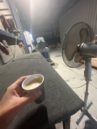 A person's hand is holding a small paper cup of coffee on the edge of a dark, textured surface. In the background, a metal fan with a black mesh grille is positioned to the right, and various industrial equipment and machinery are visible. The environment appears to be a workshop or factory setting, possibly in the city of Bogucin, Poland, given the context. The lighting is dim, suggesting an indoor space with artificial illumination. There are no other people clearly visible in the frame.