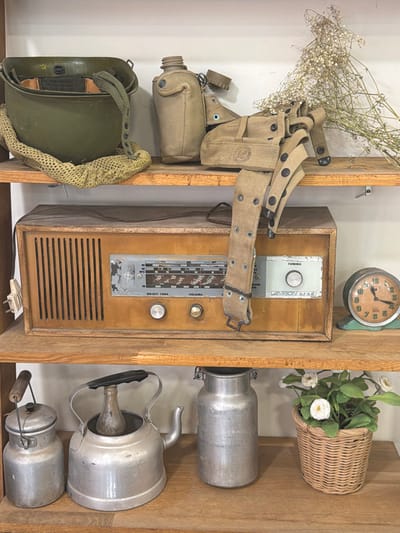 A rustic wooden shelf displays an eclectic assortment of items. On the left, a military helmet and web gear with canteen sit beside an old radio. The radio has a wood frame with a silver tuner and speaker grill. A clock is above the radio. To the right of the radio, a woven basket holding daisies and three aluminum containers occupy the shelf.