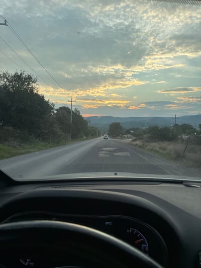 The image shows a view from inside a vehicle, likely the driver's perspective, driving on a paved road. The road stretches ahead into the distance, seemingly ascending a slight incline. Trees and vegetation line both sides of the road. In the distance, a small white vehicle is visible on the road. The sky is partially cloudy, with hints of sunset colors in the clouds. A power line runs alongside the road. The dashboard and steering wheel of the vehicle are visible, including gauges and controls. The temperature gauge shows approximately 20 degrees Celsius. The general location is consistent with Santiago Tlautla, Mexico, given the landscape and road characteristics.