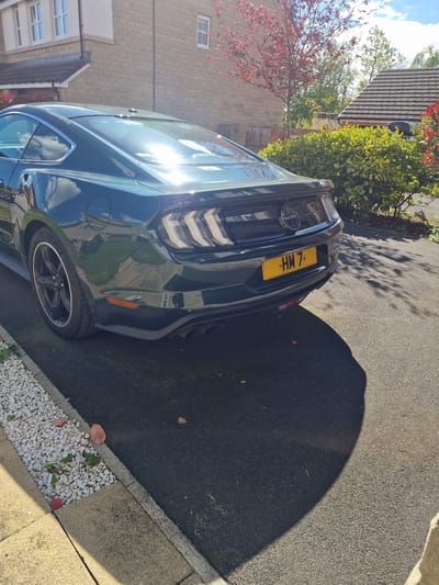 The image shows a dark green Ford Mustang parked on an asphalt driveway in Pity Me, United Kingdom. A house with a red-tiled roof and beige brick walls is visible in the background. The car's license plate is yellow with the black characters "HM 7". A bush and a tree with red leaves are also present. The edge of the driveway is lined with small white stones.
