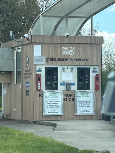 The image displays a standalone, automated fuel payment kiosk situated outdoors in Fleury-les-Aubrais, France. The kiosk features a wood-paneled exterior and is protected by an arched, translucent canopy supported by a metal frame. The structure rests on a concrete base, with a patch of green grass visible in the immediate foreground, suggesting it is located adjacent to a paved area or road.
The front face of the kiosk is dominated by two recessed payment/dispensing control units, each containing a screen, card reader, and what appears to be a cash acceptance slot or barcode scanner. These units are flanked by red emergency call points or intercoms. Above the central payment area, a prominent sign states "RÈGLEMENT EN ESPÈCES" (Payment in Cash), accompanied by symbols for 24/7 operation, credit card (CB), and a camera icon. To the left of the payment units, another vertical sign reads "ICI J'ACHÈTE MON CARBURANT MOINS CHER !" (Here I buy my cheaper fuel!), with logos for AdBlue and GPL (LPG) below it.
Below the payment units, two identical framed signs announce, "VOS BORNES À BILLETS SONT ACCESSIBLES" (Your ticket terminals are accessible) with operating hours listed as "Du Lundi au Samedi de 8h00 à 20h00 et les dimanches de 10h00 à 18h00" (Monday to Saturday from 8:00 to 20:00 and Sundays from 10:00 to 18:00), followed by "Fermé les jours fériés" (Closed on public holidays). Several white papers with additional information are taped to the kiosk facade.
Security infrastructure is visible, with at least two white dome-shaped surveillance cameras mounted high on the kiosk, one on the front-right corner and another on the left side, extending from a grey conduit. Padlocks are visible securing access panels above the payment terminals. The background features sparse-leafed trees under a partly cloudy blue sky. A distinct reflection in the lower foreground indicates the photograph was taken from within a vehicle. No individuals or active transactions are depicted.