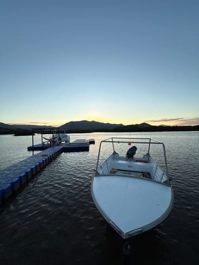 This image captures a waterfront scene at either sunrise or sunset in Dumbéa, France. The sky presents a gradient from light blue overhead to a bright yellow-orange glow at the horizon. A range of dark, vegetated hills or mountains is silhouetted against this light. In the immediate foreground, the bow of a white, open-hull boat, likely an aluminum skiff, occupies the right side of the frame, facing the viewer. It features a dark outboard motor at the stern and a metal frame extending overhead. To the left, a blue modular plastic floating dock extends into the midground. At the far end of this dock, a second, smaller white boat or structure is moored, partially obscured, adjacent to a wooden pergola-like structure containing a visible hammock. The water is dark and exhibits surface ripples. No individuals, specific actions, or interactions are depicted.