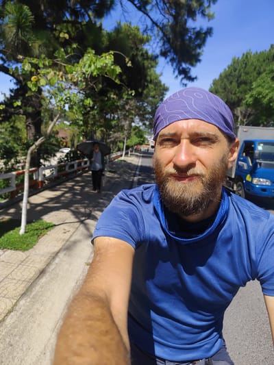 A selfie taken outdoors in Thành Phố Đà Lạt, Vietnam, shows a man with a beard and a blue bandana. He is wearing a blue shirt and has a blue neck gaiter. The photo is taken on a road next to a sidewalk with a low white and orange fence. An elderly woman carrying a dark umbrella walks away in the distance on the sidewalk. A small blue truck is parked on the road behind the man. Trees line both sides of the road, and the sky is clear and blue.