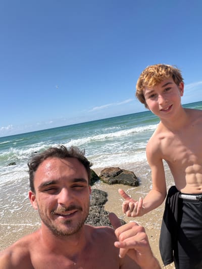 An image captured on a sunny day at a sandy beach in Beit Yanai, Israel, features two shirtless males posing in what appears to be a selfie. The foreground shows an adult male, approximately 20-30 years old, with short dark curly hair, a beard, and a mustache, smiling towards the camera with partially closed eyes. He wears a white earbud in his right ear, and his right hand is visible in the lower left corner making a gesture with his thumb and index finger extended. To his right and slightly behind him stands a younger male, appearing to be a pre-teen or early teenager, with light brown curly hair, also smiling towards the camera. He is wearing black mesh shorts with a white waistband and has his right hand raised, giving a thumbs-up gesture.
The background consists of the Mediterranean Sea with moderate turquoise waves breaking on the shore, which is composed of light brown sand and dark, algae-covered rocks. The sky is clear and bright blue, with minimal high-altitude cloud formations. A small white kite or similar airborne object is visible high in the sky in the upper left portion of the frame.