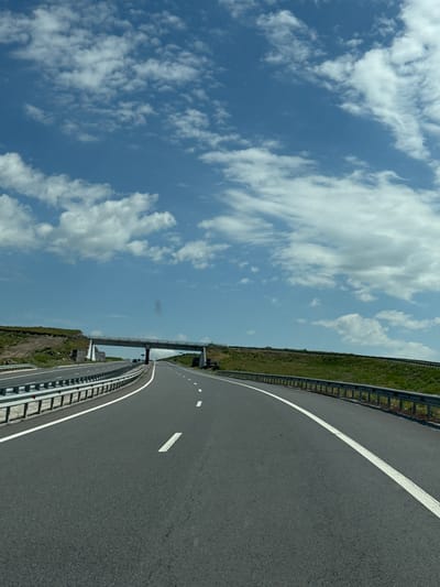 An eye-level, vertical shot captures a highway under a vast expanse of blue sky dotted with fluffy white clouds. The road, paved with dark asphalt, is divided by a white dashed line and a solid white line on the right side. A metal guardrail runs along the edge of the road. In the distance, an overpass crosses the highway, supported by concrete pillars. Green grassy hills rise on either side of the road, with some exposed soil on the left. The scene appears to be a rural highway, possibly in or around Lanjik, Armenia, given the context. There are no people or vehicles clearly visible in the immediate foreground or mid-ground. The lighting suggests a bright, sunny day.