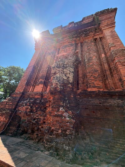 A low-angle shot captures a weathered brick tower, likely a Champa ruin, under a bright blue sky with strong sunbeams. The tower, constructed from reddish-brown bricks, shows signs of age and erosion, with patches of moss and discoloration. Vertical indentations are visible on its surface, adding to its ancient appearance. The sun is positioned in the upper left corner, casting a lens flare across the image and illuminating the top portion of the tower. To the left, the lush green foliage of a tree is visible against the sky. The ground in the foreground is paved with stone tiles, casting a long shadow from the tower. This scene is likely located at the Thap Nhan Cham Towers, a historical landmark in Tuy Hoa city, Phu Yen province, Vietnam, a site rich in Champa heritage.