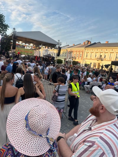 The image captures an outdoor event, likely a festival or concert, taking place in the Market Square of Nowy Sącz, Poland. A large stage is set up at the far end of the square, with a crowd of people gathered in front of it. The architecture of the surrounding buildings, typical of historic European squares, is visible in the background, featuring ornate facades and tiled roofs. Many people are standing and milling around, some appearing to be engaged in conversation. In the foreground, the back of a person wearing a wide-brimmed straw hat and a colorful, patterned top is visible. To the right of this person, another individual in a striped shirt and a white baseball cap is holding a water bottle. The atmosphere appears to be relaxed and social, with people enjoying the event under a clear sky.