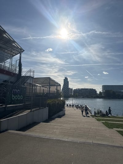 The image captures a sunny day at the Rhine riverfront in Weil am Rhein, Germany. In the foreground, a paved walkway with metal gratings leads down towards the river. To the left, a modern building with a red lower level and glass facade features an outdoor staircase. A sign for "mai boba" is visible on a fence in front of this building. Further along the riverbank, outdoor seating areas with tables and chairs are set up. Several people are seen walking and sitting by the water. Across the river, the cityscape of Basel, Switzerland, is visible, characterized by various buildings, including a distinctive tall, angular skyscraper. The sky is blue with scattered clouds and contrails from airplanes, and the sun is shining brightly, creating lens flare effects.