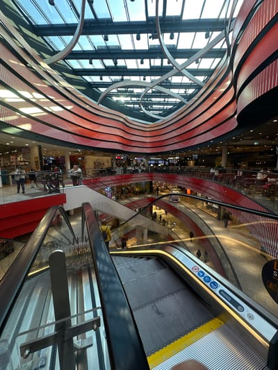 This media file captures the interior of a modern, multi-level shopping mall, viewed from an elevated position and rotated 90 degrees clockwise. The scene is dominated by contemporary architecture featuring curved lines and a striking red and black color scheme.

On the left side of the rotated image, large glass windows allow natural light to illuminate the space, framed by dark structural beams. Above and curving across this section are wavy, layered architectural panels in red and black, creating a dynamic ceiling or wall design. Metallic, circular decorative elements are also visible near the windows.

On the right side of the image, several escalators diagonally traverse the different levels of the mall. The foreground features a large escalator with silver handrails, gray ridged steps, and a bright yellow edge at the bottom. Various safety and warning symbols are visible on its side panel, including icons for no bare feet, holding the handrail, no running, no pushing carts, and a dog symbol. A yellow and black circular sign with text (partially visible as "S" or "L" and icons) is also present.

Below and between the escalators, multiple levels of the mall are visible, bustling with activity. Numerous people are scattered across these floors, appearing as shoppers or visitors. Storefronts and seating areas for cafes or restaurants line the levels, with visible text including "Burger BRO", "Kitchen", and "SATATTIO". Other signs indicate "WC" with an arrow and a level marker "2.OG". The overall impression is one of a busy, well-lit commercial space, likely during the daytime due to the bright light from the windows. The contemporary design elements suggest a recent construction or renovation. The location is specified as Bielefeld, Germany.