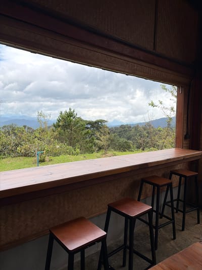 This image depicts an indoor scene looking out through a wide, wooden-framed window onto a lush, mountainous landscape in Ban Khun Wang, Thailand. In the foreground, a wooden counter stretches across the frame, with three dark-legged, wooden-topped bar stools positioned in front of it. The wall behind the counter is covered in a woven bamboo pattern. The window reveals a vista of rolling hills covered in dense green vegetation, with a cloudy sky overhead. A single pine tree stands out amongst the greenery, and there are hints of human presence, such as a blue and yellow faucet pipe visible on the grassy slope outside. The lighting suggests it is daytime, possibly overcast. There are no people visible in the scene.