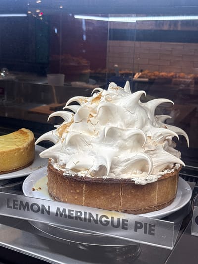 In a display case, a lemon meringue pie sits on a white plate. A label reading "LEMON MERINGUE PIE" is visible. The pie features a tall, swirled meringue topping. The photo may have been taken in Milnerton, South Africa.