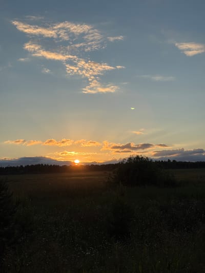The image depicts a sunset over a rural landscape in Balakli, Russia. The sun is low on the horizon, casting a warm orange glow across the sky. Wispy clouds are illuminated by the setting sun, creating a beautiful contrast with the deep blue sky. A dark line of trees stretches across the middle ground, with a field of grass in the foreground. The overall mood is serene and peaceful. The time of day is sunset, and the weather appears clear. There is no visible text in the image.