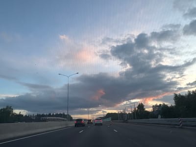 The image shows a view from a vehicle driving on a multi-lane highway, likely in the vicinity of Аист Снт, Russia. The sky is filled with dramatic clouds, with patches of pink and orange light suggesting a sunset or sunrise. On the right side of the road, a barrier with red and white striped poles is visible, possibly indicating ongoing construction or a safety zone. Several vehicles are on the highway, including a white car in the center lane and a darker car in the left lane. Trees and foliage line the side of the road in the distance. A tall lamppost is also present on the left side of the highway. The road itself is paved and marked with white lane lines.