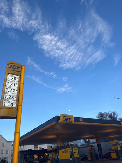 A JET gas station displays fuel prices under a bright blue sky with scattered clouds. Diesel is priced at 1.709, Super E10 at 1.719, Super at 1.779, Super+ at 1.869, and AdBlue at 1.109. The station's canopy, also marked with the JET logo, covers the pumps, with AdBlue also prominently displayed. A hint of urban architecture is visible in the background.
