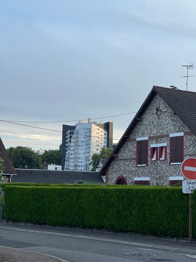 The image depicts a street scene in Maromme, France, focusing on residential buildings. In the foreground, a portion of a stone-clad house with dark brown shutters and red trim is visible. Adjacent to it is a neatly trimmed green hedge bordering the property. A traffic sign with a red circle and a white horizontal bar, indicating prohibition, is mounted on a pole to the right of the hedge. Below it, another white sign with the word "SAUF" (except) and a bicycle symbol is also present, suggesting an exception for cyclists. Beyond the hedge and the houses, a tall, modern white and grey apartment building stands prominently in the background, contrasting with the older architecture. Trees and other smaller buildings are scattered between the foreground and the apartment block. The sky is overcast with a pale blue hue, indicating early morning or late afternoon light. The street itself appears to be paved, with a sidewalk and a manhole cover visible. There are no people or animals present in the frame.