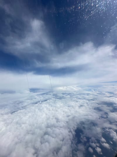 The image depicts a view from an airplane window, looking down at a vast expanse of clouds. The sky above is a deep blue, with lighter, wispy clouds interspersed with darker, more textured ones. The clouds below form a thick, undulating carpet, with patches of deep blue sky visible between them. The scene is characterized by the interplay of light and shadow across the cloud formations, creating a sense of depth and dimension. There are visible scratches and smudges on the airplane window, which distort the view slightly. There are no people, animals, or man-made structures visible in the image, nor any indication of the city of Yur'evo, Russia. The environment is purely atmospheric, showcasing the natural beauty of the sky from a high altitude.