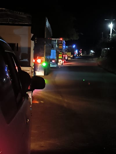 The image depicts a night scene on a road in Lhoknga, Indonesia, viewed from inside a parked or slow-moving vehicle. The perspective is forward and slightly to the right, showing a line of large trucks and other vehicles.
In the foreground on the left, elements of the capturing vehicle are visible, including a dark side window frame, the exterior side mirror, and a portion of the car's body. Reflections of various lights, predominantly red and green, scatter across the car's dark surface.
The midground is filled with a queue of large trucks. The closest truck, positioned to the right of the foreground car, displays prominent red taillights and a bright green signal light. Below its rear, the word "JAYA" is visible in white letters. The road surface below the trucks is dark but reflects the vehicle lights, creating colorful streaks.
Further down the road, more vehicles are discernible, characterized by their red, blue, and yellow lights, suggesting a continued line of traffic or parked heavy vehicles. On the far right, a bright street light pole illuminates a section of the road and the dark, obscured foliage. The overall setting is dark, emphasizing the artificial lights from the vehicles and the street. The scene conveys a sense of night travel, possibly involving a traffic jam, a waiting area, or a convoy.