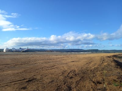 The image depicts an outdoor scene at an airstrip located in Santa Juana, Chile, under a bright blue sky with scattered white cumulus clouds. The foreground consists of dry, reddish-brown earth with numerous discernible vehicle tire tracks and minimal sparse green vegetation along the right edge.

In the mid-ground, a dark gray paved runway extends horizontally across the scene. A single-engine, propeller-driven aircraft, predominantly white with red markings on its tail section, is stationary on the paved surface, positioned to the left of the frame's center. Its propeller blades are static. To the immediate left of the aircraft, on the same paved surface, two white, dome-shaped structures are present; one is a larger, prominent circular structure, and the other is smaller and partially obscured behind it.

Beyond the paved runway, the mid-ground features a vast expanse of disturbed, light brown earth, with several dark, irregular piles of material visible along the distant horizon line. The background is dominated by a continuous, dark green forested area or low mountain range under the clear sky. No individuals are visible within the frame, and no active movement or interaction is depicted.