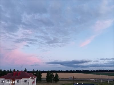 The image depicts a rural landscape under a partly cloudy sky, with a distinct pink hue to the clouds, suggesting either dawn or dusk. In the foreground, a two-story building with a red-tiled roof is visible on the left side of the frame. The building has white walls and several windows. To the right of the building is a vast expanse of agricultural fields, some appearing golden brown, possibly indicating harvested crops or ripe grain, while others are a vibrant green. A dark line of trees forms a dense border between the fields and the horizon. The sky is a mix of blues and greys, with the aforementioned pink clouds creating a dramatic effect. Several indistinct structures, possibly utility poles or small buildings, are scattered across the landscape. The overall impression is one of a tranquil, rural setting in Улукаўскі сельскі Савет, Belarus. There are no people visible in the image.