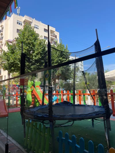 The image shows an outdoor children's play area with a trampoline in the foreground. The trampoline has a black safety net and is surrounded by a colorful plastic fence. Behind the trampoline, there is a red and green slide, and further back, a tall, cream-colored apartment building. Trees with green foliage are visible in front of the building. The sky is clear and blue. The scene appears to be in a city, and the presence of a building, trees, and an outdoor play area suggests an urban environment. The flags at the top left of the image could indicate a location in Bashkia Durrës, Albania, as flags are often flown at businesses or public spaces in such areas.