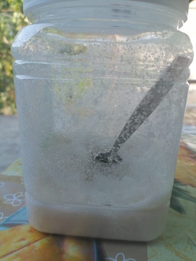 The image shows a clear plastic jar filled with a white, granular substance that appears to be sugar or salt. The jar is partially filled, with a layer of the granular material at the bottom, and the rest of the jar filled with smaller crystals and some liquid. A metal spoon is submerged in the contents of the jar, resting at an angle. The jar is placed on a colorful, patterned surface, possibly a tablecloth or placemat. In the background, out of focus, is greenery that suggests an outdoor setting. The lighting indicates it's daytime, with sunlight filtering through the contents of the jar and illuminating the crystals. The scene appears to be set in Gumbaz, Uzbekistan, as per the provided location context. There are no people visible in the image. The overall impression is one of a common household item, possibly used for storage or preparation of food or ingredients. The focus is entirely on the contents of the jar and the spoon within it, with the background providing only general environmental context.