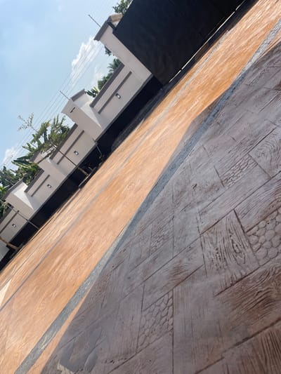 The scene depicts an exterior ground-level view of a property entrance or driveway in Ogbunu, Nigeria. The ground surface is paved with stamped concrete, featuring two distinct sections. The left and upper portion of the paving is a reddish-brown color with a textured finish. The right and lower portion is grayish-brown, imprinted with multiple patterns resembling wood grain and scale-like textures. A thin, dark grey strip runs diagonally, separating these two paved areas. In the background, a white perimeter wall with dark grey accents is visible, featuring several integrated rectangular light fixtures. Above the wall, green foliage from trees and shrubs is present, along with multiple parallel wires, indicative of utility or security infrastructure. A large, dark-colored gate is partially visible behind the wall on the right side. The sky above is clear blue with sparse white clouds. The lighting suggests a bright, sunny day, with shadows cast across the paved surface. No individuals, actions, or interactions are observed in the frame.