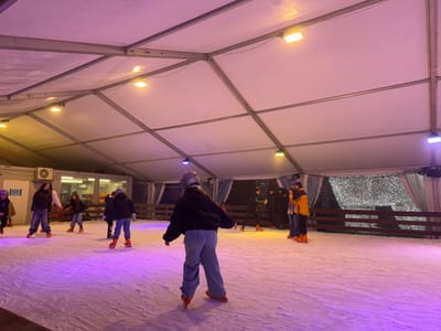 Several people are ice skating inside a large tent-like structure. The tent has a white roof supported by metal beams and is illuminated by overhead lights. Some skaters wear helmets, and all are wearing ice skates. A wooden fence surrounds the ice rink, with a backdrop of trees and lights visible behind it. An air conditioning unit is visible on the side of a building adjacent to the rink.