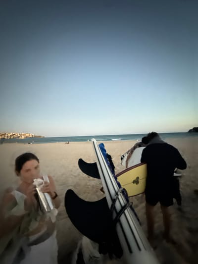 The image captures a twilight scene at Bondi Beach, Australia. In the immediate foreground on the left, a female individual in a light-colored top is observed drinking from a silver metallic shaker bottle, holding a white crumpled object in her left hand. To her right, two surfboards are present: a prominent white surfboard with black longitudinal stripes and visible black fins leans towards the viewer, partially obscuring a yellow surfboard with a black fin and dark symbol. In the mid-ground, a silhouetted male figure in dark clothing walks away from the camera, carrying a white surfboard under his right arm. The background features the sandy beach, with the ocean and small waves visible further out. On the far left horizon, the illuminated buildings of a city or town are visible on a distant headland. Several other indistinct figures are scattered on the beach closer to the waterline. The sky exhibits a gradient from dark blue overhead to a lighter blue-grey near the horizon, indicative of dusk or dawn.