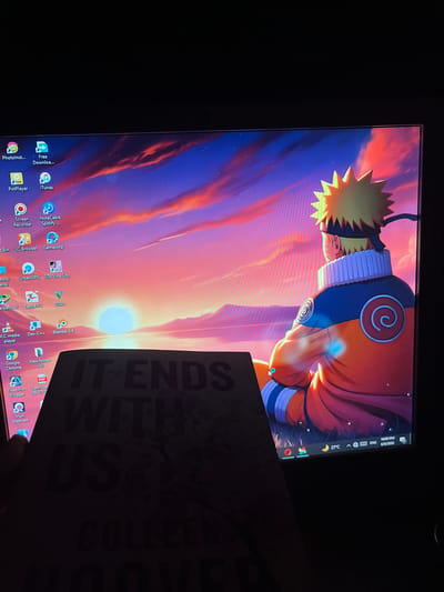 The image displays a computer monitor screen partially obscured by a book, held by a human hand, in a dimly lit environment located in the city of Kabal, Pakistan.
The monitor's display features a desktop background depicting the anime character Naruto Uzumaki from the back, looking towards a sunset or sunrise over a landscape. The sky exhibits vibrant orange, red, and purple hues with a prominent bright light source on the horizon. Naruto is depicted in an orange and blue uniform with a white-collared jacket and a black headband, showing a red spiral emblem on his left shoulder.
Various application icons are visible on the desktop, including: PhotoInst..., Free Downloa..., PotPlayer, iTunes, Recycle Bin, Screen Recorder, NoteCable Spotify..., UC Browser, Gameloop, Control Panel, UrbanVPN, GTA Vice City, BlueStacks X, Zapya PC, GTA5, VLC media player, Dev-C++, Blender 3.4, Google Chrome, New folder (4), VideoProc Vlogger, PDF, CamScanner 09-27-2..., and Irun Webcam.
The Windows taskbar is partially visible at the bottom right, displaying a moon icon, 27ยฐC, language set to ENG, the time as 10:00 PM, and the date 6/6/2026.
A book, identified by the visible text "IT ENDS WITH US" and "COLLEEN" (presumably for Colleen Hoover), covers the lower portion of the monitor screen. The book's cover appears dark. A human thumb is visible on the left side, holding the book against the monitor. The room environment outside the illuminated screen appears dark.