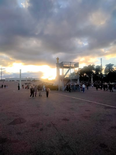 The image presents a wide-angle view of a vast, paved outdoor plaza within the Sanctuary of Fátima, Portugal, under an overcast sky during late afternoon or early evening. A low sun breaks through the clouds on the horizon, creating a bright, warm backlight that silhouettes many elements and casts long shadows.
In the left mid-ground, a tall, dark concrete crucifix with a minimalist design stands prominently. Further to the left and in the background, a long, white, modern building, likely part of the Sanctuary complex, is visible beneath the bright horizon.
In the right mid-ground, a multi-level, open-framework metallic structure, possibly a temporary platform or viewing area, is erected. A golden statue of a religious figure stands atop a white pedestal on the upper left side of this structure. Adjacent to the right of this structure, a dense cluster of trees is visible, along with a tall, slender pole. Metal barrier fencing is present around the base of the metallic structure.
The foreground is a dark, paved surface, showing minor cracks and textural variations. A distinct white line is painted on the ground, extending from the lower right towards the center.
Numerous individuals are dispersed throughout the plaza. In the immediate foreground, a group of approximately five people are walking from left to right, away from the viewer. Dozens of other individuals and smaller groups are scattered across the mid-ground and background, walking or congregating. Some individuals are visible near the metallic structure, behind the barriers, and in the distant areas of the plaza. All observable actions are consistent with walking or standing in a public outdoor space.