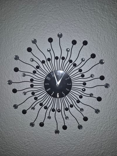 A decorative wall clock hangs on a textured, light-colored wall. The clock has a black face with white Roman numerals and a sunburst design made of thin black metal rods. Each rod terminates in either a small black sphere or a clear, faceted bead. The clock's hands indicate the time is approximately 11:46. The background is a uniformly textured wall, likely interior. The image is taken from a medium distance, showing the entire clock and a portion of the surrounding wall. There are no people or animals present.