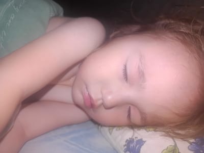 The image shows a close-up of a young child sleeping peacefully. The child is lying on their side, with their head resting on a pillow. Their eyes are closed, and their lips are slightly parted, suggesting they are in a deep sleep. The child's skin appears soft and fair, and their hair is light brown and somewhat disheveled. They are wearing a light-colored garment, possibly a onesie or pajama.
The setting appears to be a bedroom, indicated by the presence of a pillow and what looks like a blanket or duvet in the background. The lighting is dim, suggesting it might be nighttime or early morning. There is a green blanket with some faint white text or pattern on it visible in the upper left corner of the image. The pillow the child is resting on has a floral pattern with blue and green elements. The overall mood of the image is one of tranquility and innocence. There are no external cues to definitively determine the exact time of day or weather conditions. The child's expression is serene, conveying a sense of comfort and security. The location context provided is Esik, Kazakhstan.