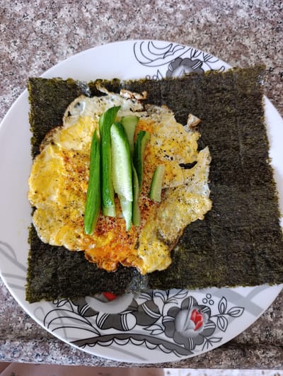 A fried egg and cucumber slices are placed on a sheet of roasted seaweed, which rests on a white plate with a floral pattern. The plate is on a speckled granite countertop. The scene is indoors, with no visible indication of the city of Nablus, Palestinian Territories.