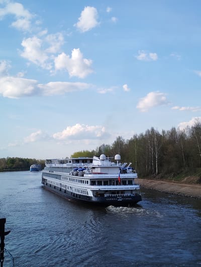 The image shows a river scene in Varvarino, Russia. A large, multi-decked cruise ship with "КНЯЖНА ВИКТОРИЯ" written on its stern is moving along a river or canal. A smaller blue and white boat is visible in the distance. The water is dark and rippled, indicating movement. The riverbanks are lined with trees, mostly leafless, suggesting an early spring or late autumn season. The sky is bright blue with scattered white clouds.