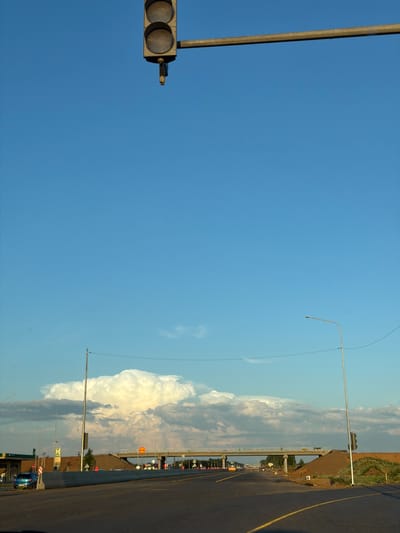 The image captures a roadside scene under a clear blue sky with a dramatic cloud formation in the distance. A traffic light hangs over an empty road, suggesting a controlled intersection. The road appears to be asphalt with yellow lane markings. A concrete barrier separates the lanes of traffic from the surrounding environment, which includes a grassy embankment and some construction activity. In the background, a bridge spans over the road, and streetlights are visible on either side. A blue car is parked near a building that resembles a gas station. The overall impression is one of an open road in a somewhat rural or suburban setting, possibly on the outskirts of Derzhavino, Russia.