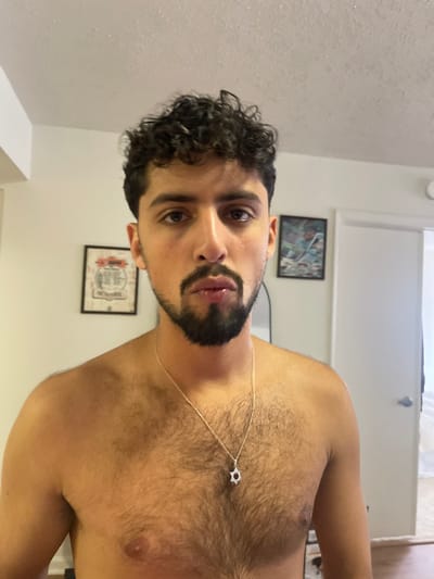 The image displays a male individual, bare-chested, with dark curly hair and a dark goatee-style beard, looking directly at the camera. He wears a silver-colored chain necklace with a Star of David pendant. His chest and abdomen are visibly hairy. The scene is an indoor residential room, with light-colored walls and a white, textured "popcorn" ceiling. On the left wall hangs a framed graphic poster with circular elements. On the right wall, a framed artwork depicts a dynamic, blue and green-toned scene. A white door, partially ajar with a silver handle, is visible on the far right, with a dark-framed object (likely a mirror) partially visible behind the subject's right shoulder. While consistent with a dwelling in Arlington, United States, the exact location within the city cannot be determined from the image.